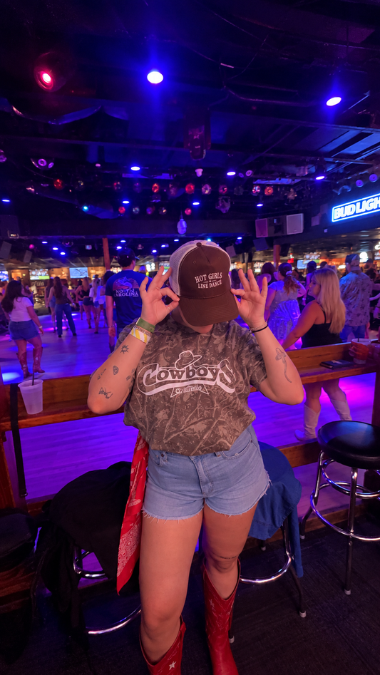 A girl standing in a nightclub wearing a brown trucker hat with 'HOT GIRLS LINE DANCE' embroidered on the front, a camouflage graphic t-shirt, denim shorts, and red cowboy boots. The background is filled with neon lighting, bar stools, and other people.