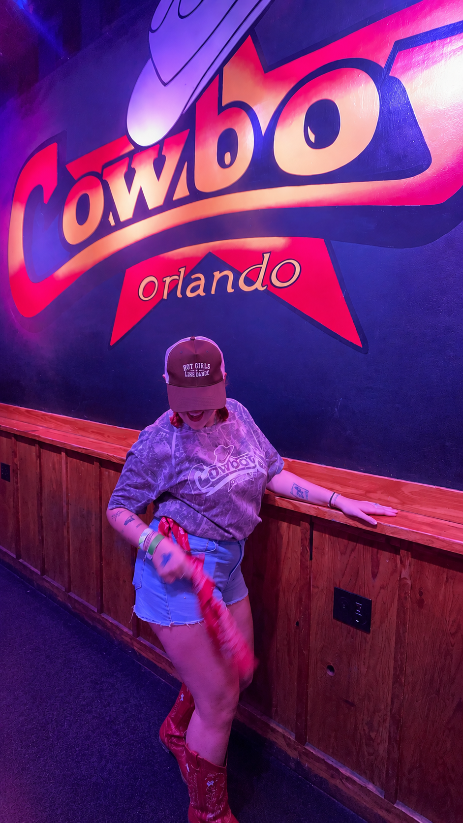 A woman posing in front of a large 'Cowboys Orlando' sign, wearing a brown cowboy hat, a gray graphic t-shirt, denim shorts, and red cowboy boots while twirling a red bandana