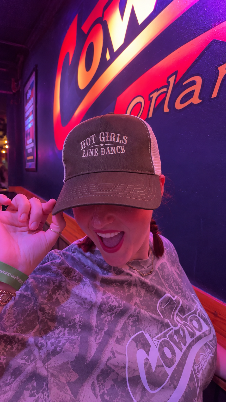 A person wearing a brown trucker hat with 'HOT GIRLS LINE DANCE' embroidered on the front, posing in front of a brightly lit bar sign with purple and orange lighting.
