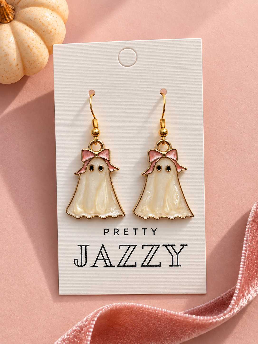 A pair of ghost-shaped drop earrings with gold hooks and pink bows, displayed on a white card that reads 'PRETTY JAZZY' against a soft pink background with decorative pumpkins and ribbon.