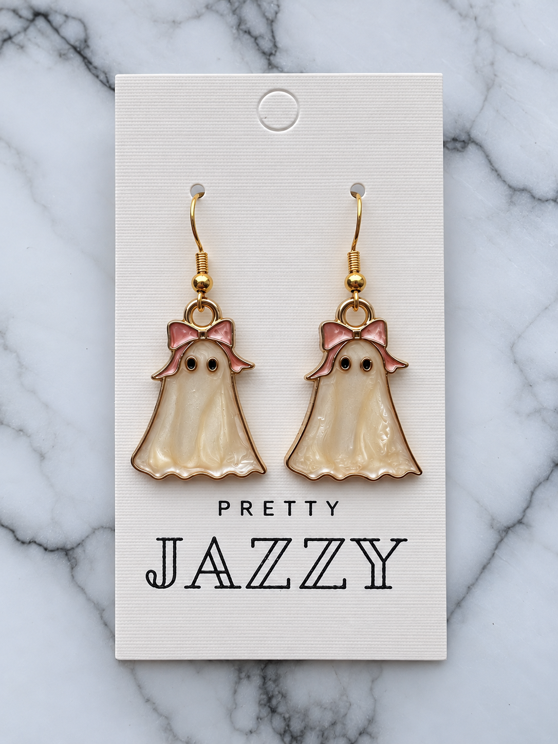 A pair of dangling ghost-shaped earrings with pearly cream enamel bodies, pink bows, and gold hooks, displayed on a white branded card over a marble surface. The card reads 'PRETTY JAZZY'.