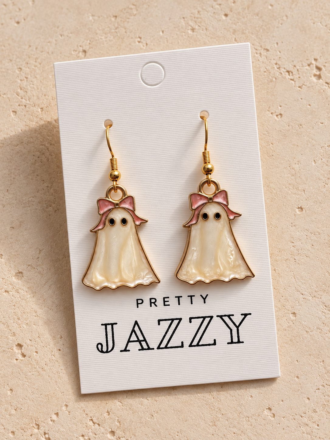 A pair of ghost-shaped dangle earrings with gold hooks, cream enamel bodies, pink bows, and small black eyes, displayed on a white card printed with the brand name 'PRETTY JAZZY' against a textured beige background.