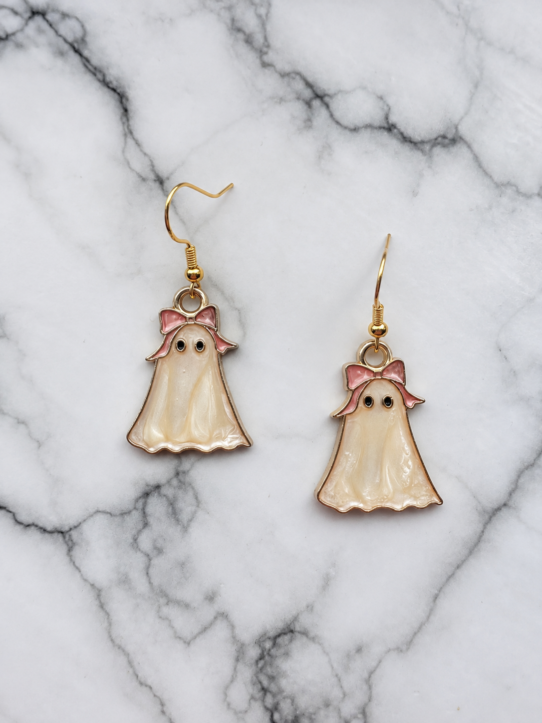 A pair of dangling ghost-shaped earrings with pearly cream enamel bodies, pink bows on gold hooks, displayed on a white and gray marble surface.
