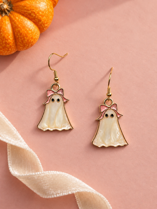 A pair of gold-tone dangle earrings - cute pearly white ghosts with pink bows on their head. Displayed on a dusty pink background with a decorative pumpkin and cream velvet ribbon.