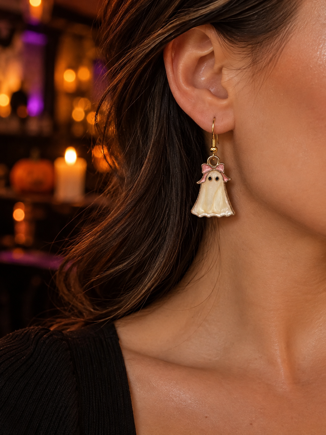 A close-up of a woman's ear wearing a dangling ghost-shaped earring. The earring has a gold-tone hook, a white pearlescent ghost body, small black eyes, and a pink bow at the top.
